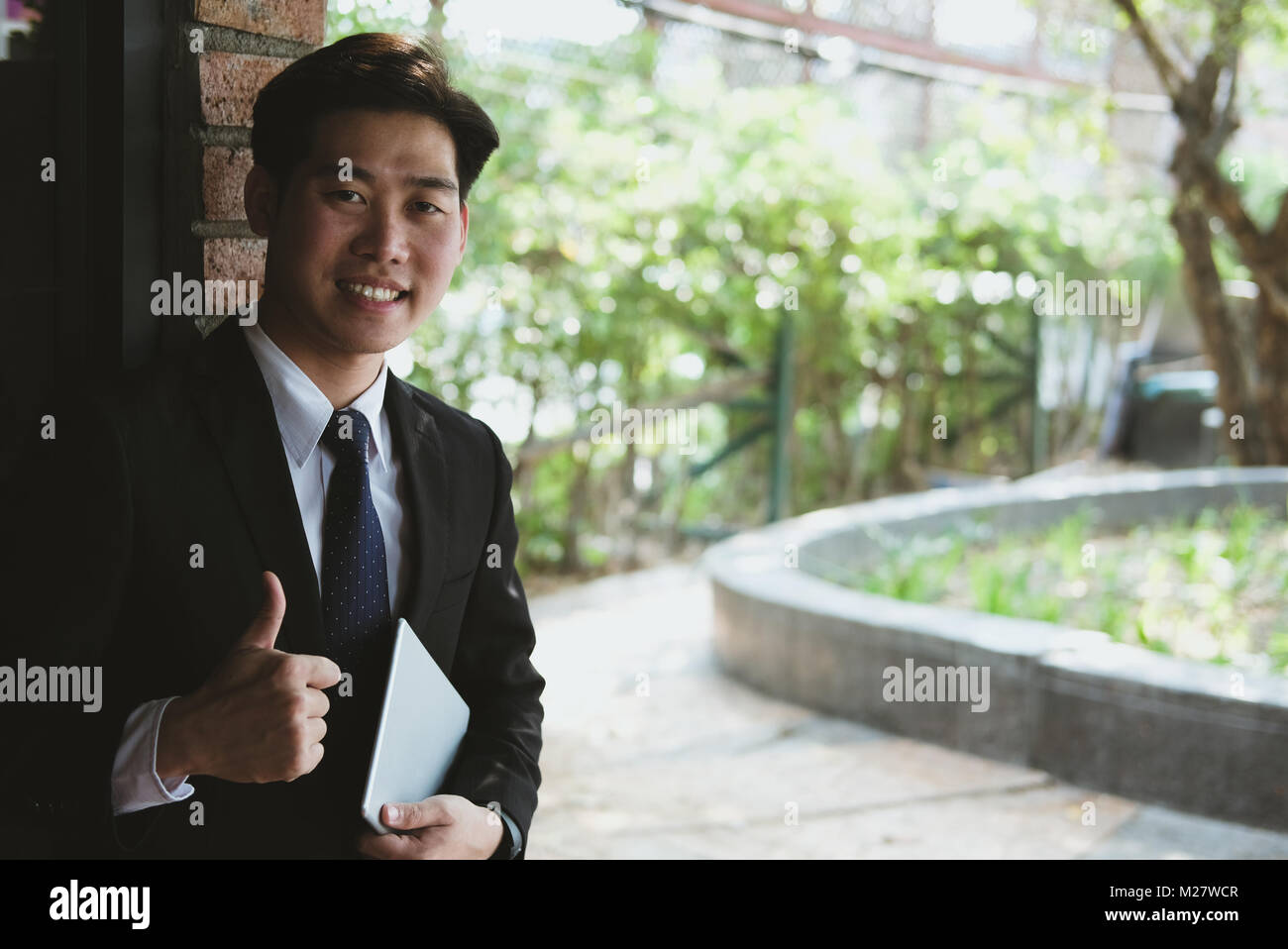 confident young businessman in suit holding digital tablet while standing outside building. asian startup man showing thumb up outdoors Stock Photo