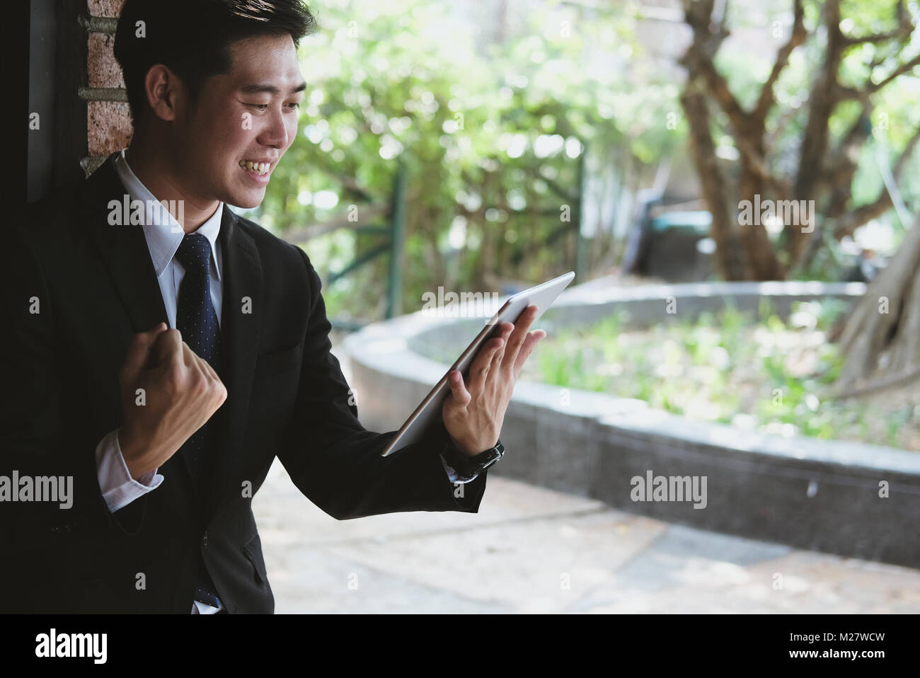 Successful business man checking tablet message & raise fist with happiness and gladness outside office building. male startup entrepreneur enjoy succ Stock Photo