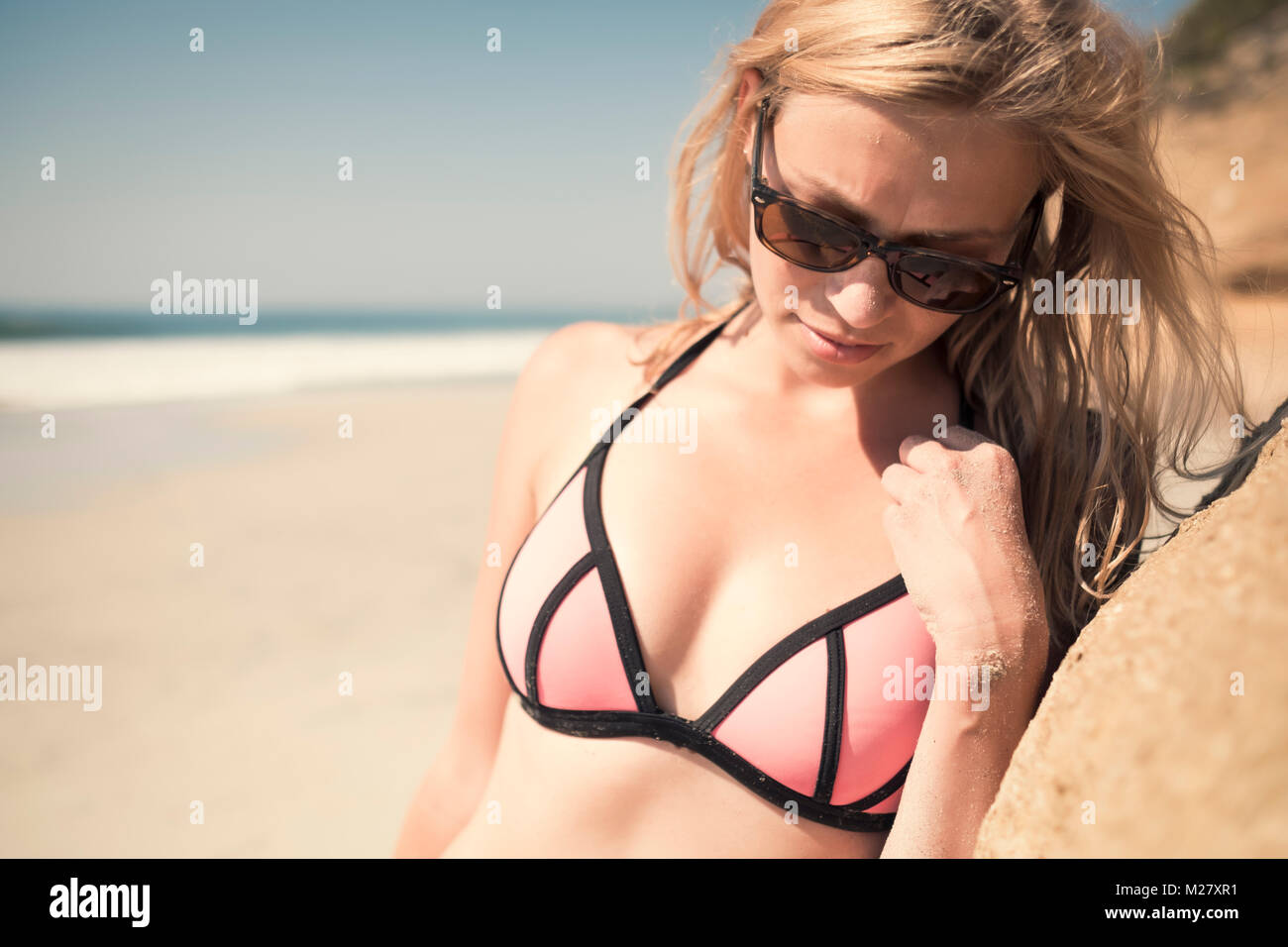 Cose up portrait of young caucasian woman in bikini, wearing sunglasses, leaning against a rock at a beach. Stock Photo