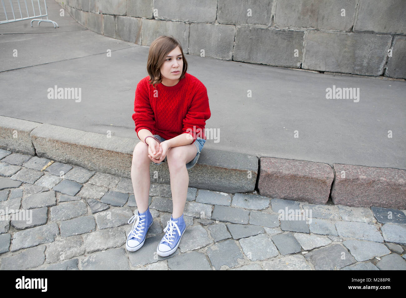 A young girl in a red wool sweater and jeans shorts is sitting on the steps Stock Photo