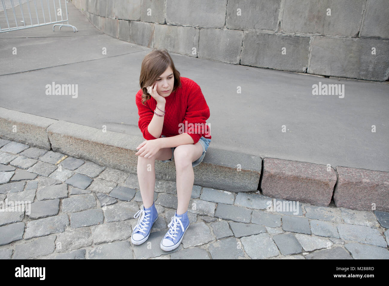 A young girl in a red wool sweater and jeans shorts is sitting on the steps Stock Photo