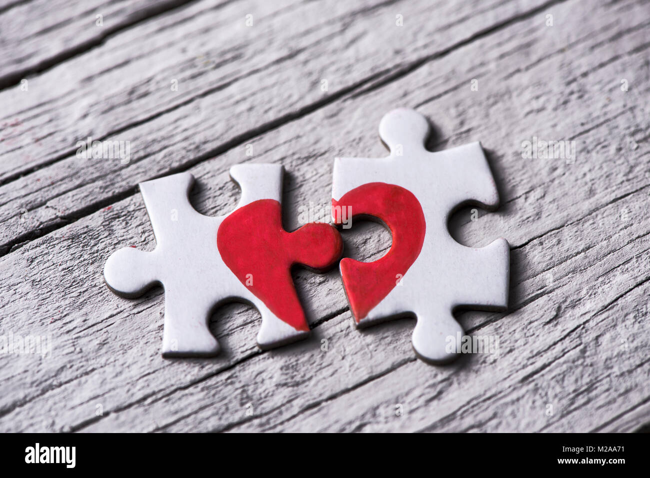 closeup of two separated pieces of a puzzle which together form a heart on a white rustic wooden surface, depicting the idea of rupture or cooperation Stock Photo