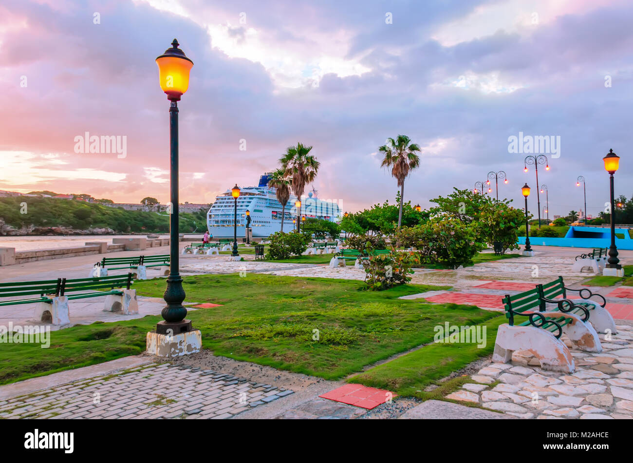 Embankment with luminous yellow light street lamps, palm trees, blue cruise liner, pink clouds, benches and green grass in morning Havana Stock Photo