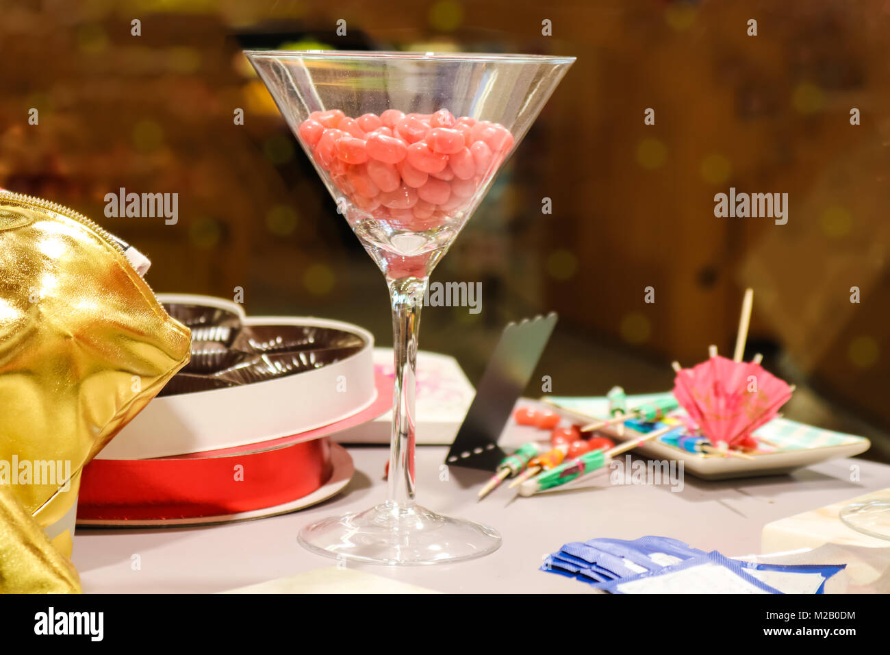 Martini glass filled with pink jellybeans on table with scattered drink umbrellas and mostly empty valentine chocolate box - selective focus Stock Photo