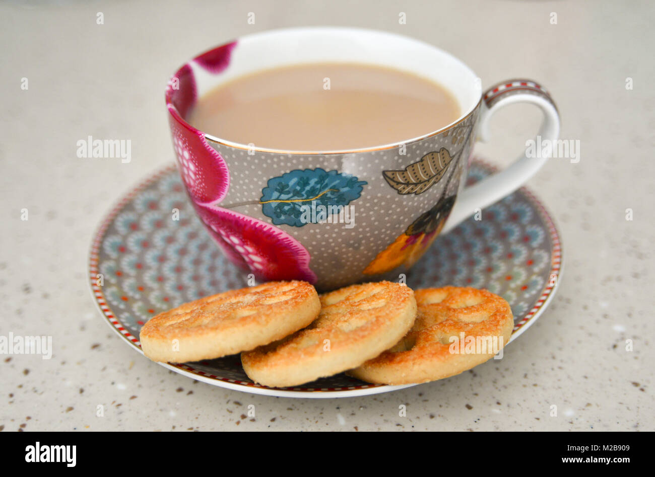 Cup of tea with milk, in a floral patterned cup and saucer with three round biscuits on a beige surface Stock Photo