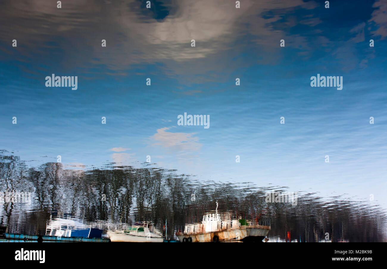 Blurry reflection of anchored old boats, trees, blue sky and clouds in river water Stock Photo