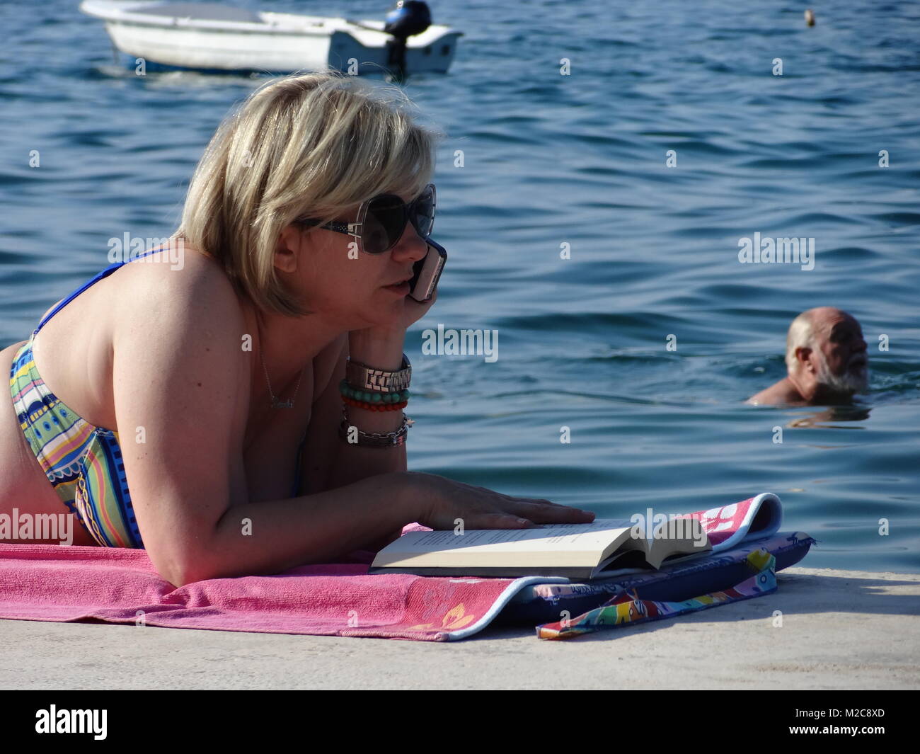Die Füße hoch legen, Sommer, Sonne und mehr - die Urlauber freuen sich auf die schönsten Zeit des Jahres. Eine Frau liest ein Buch und telefoniert am Strand. Telefonieren in der EU ist seit dem 1. Juli 2013 billiger.  Eine Frau telefoniert mit dem Handy am Strand /Beach. Stock Photo