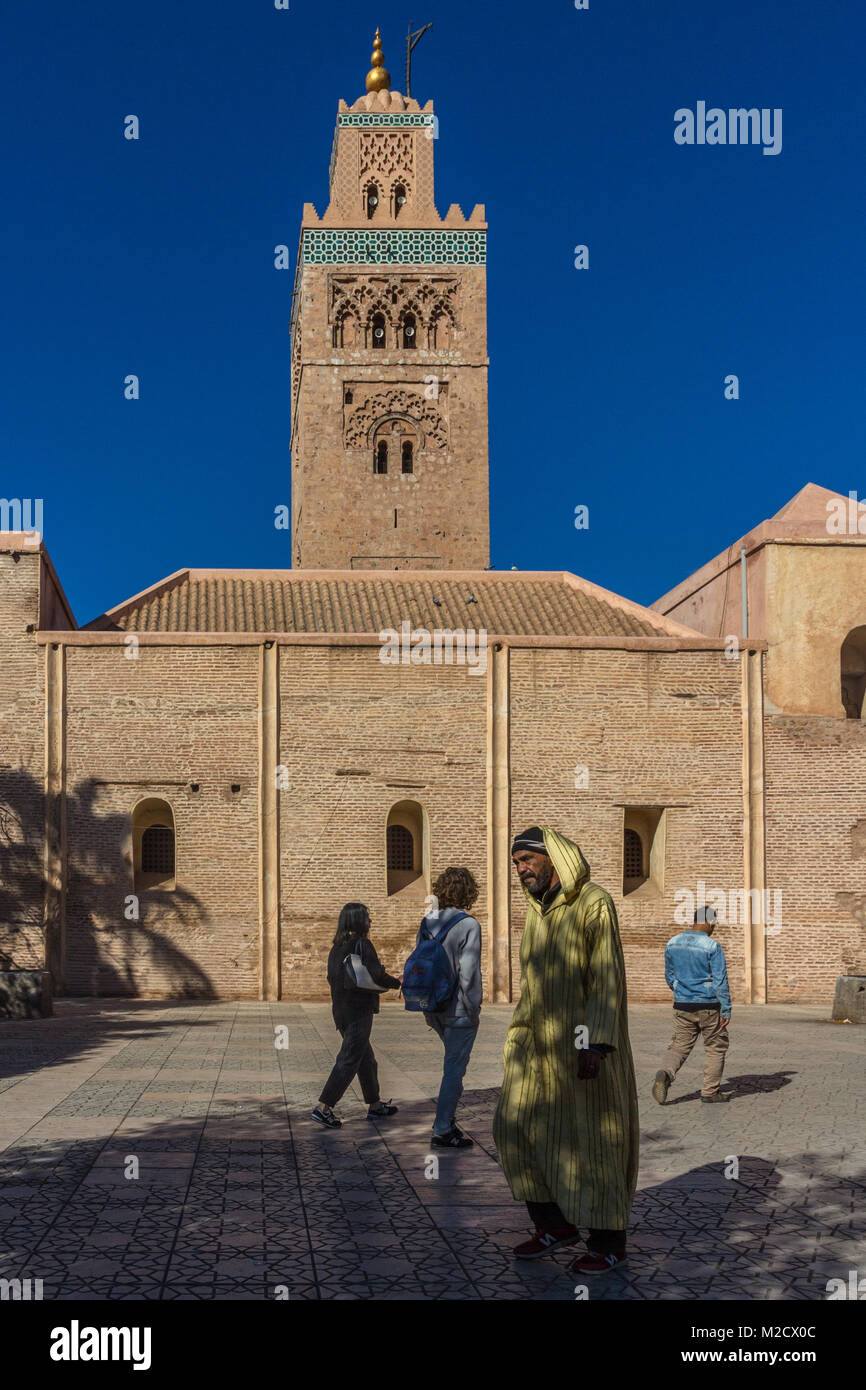 A man wearing a yellow djellaba walks by Koutoubia Mosque and Minaret, in Marrakesh, Morocco, while a group of young tourists walk the other way. Stock Photo