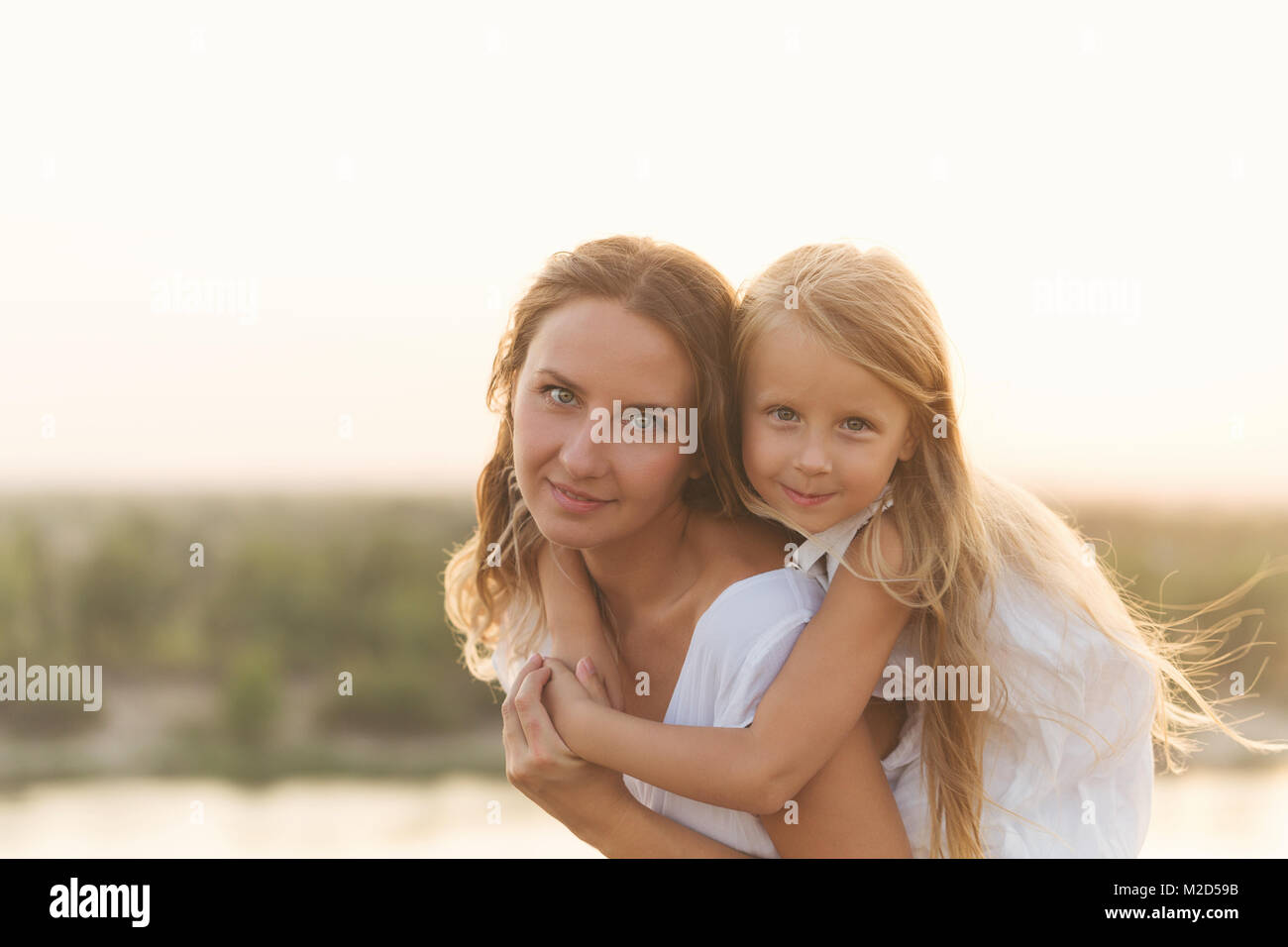 Family, mother and daughter. Girl Piggyback Daughter. Girls in white dresses. They are blondes. Family time together. Stock Photo