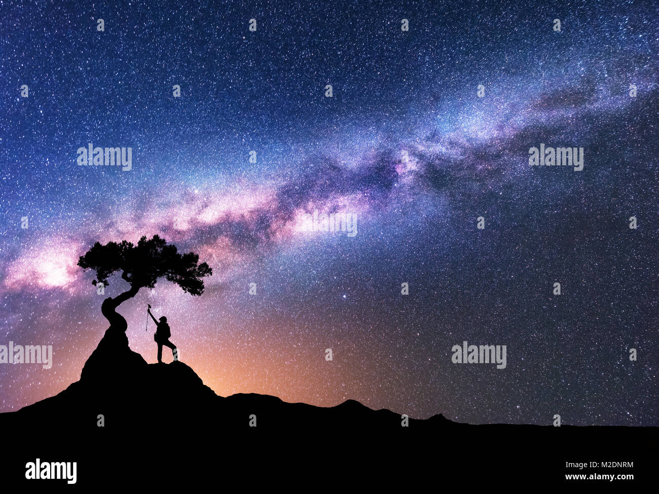 Milky Way and silhouette of woman under the tree growing from the rock on the mountain at night. Space background with starry sky, beautiful galaxy an Stock Photo