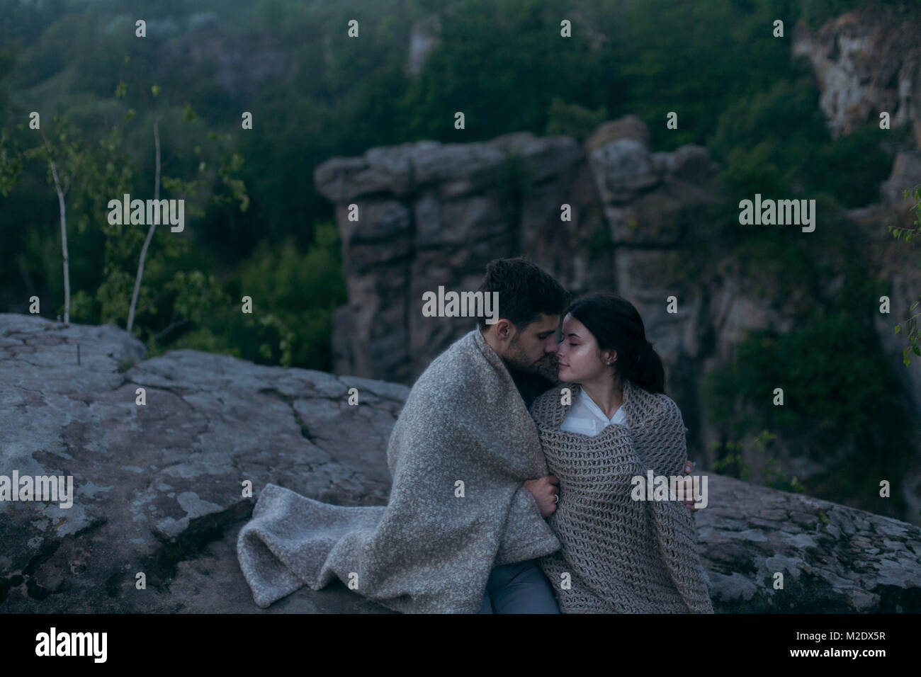 Caucasian couple sitting on rock wrapped in blanket Stock Photo