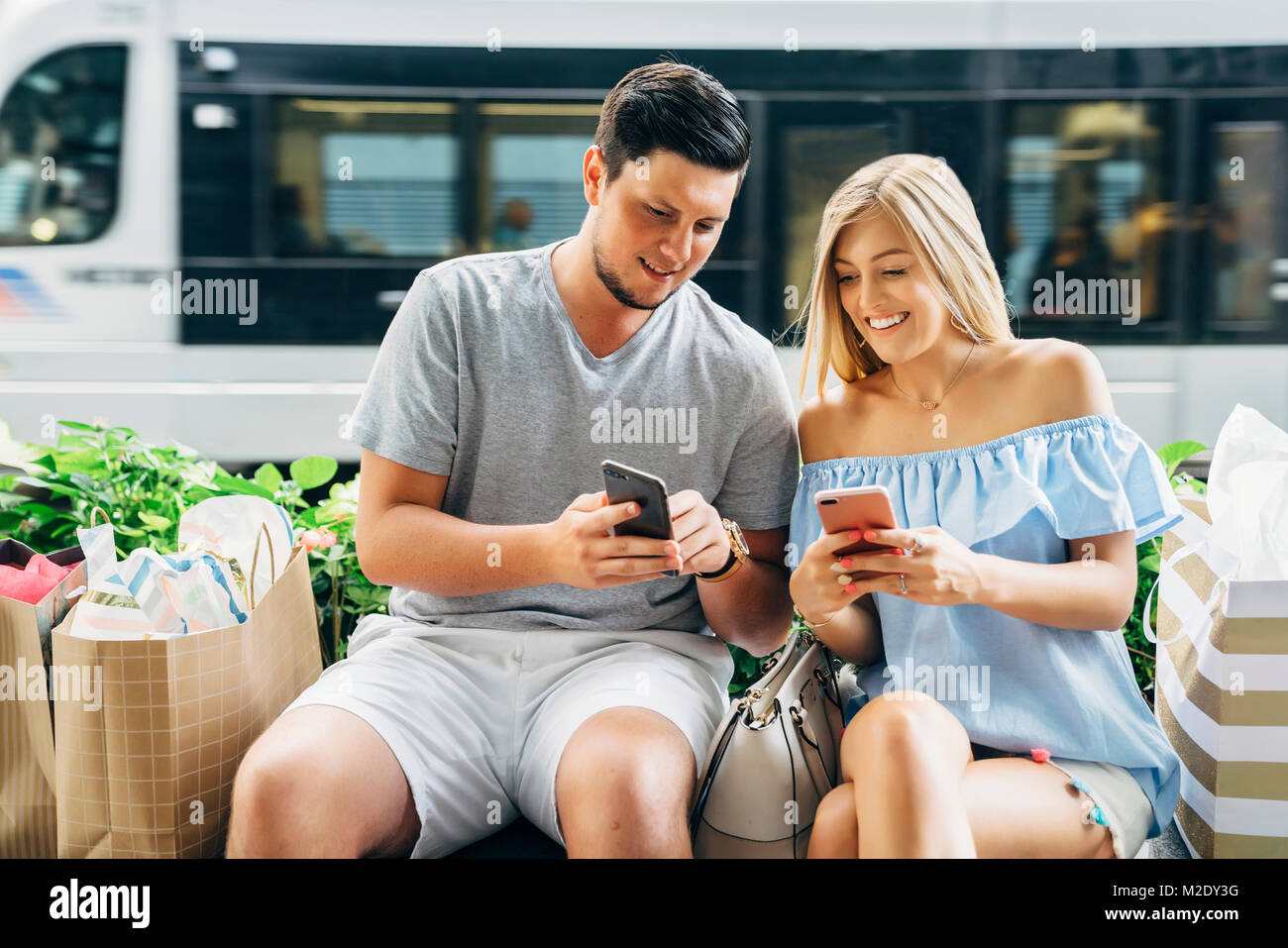 Caucasian couple with shopping bags texting on cell phones near train Stock Photo