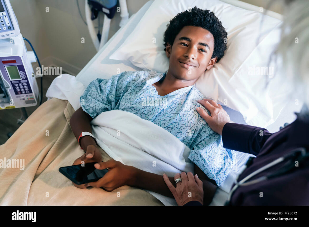 Doctor comforting boy laying in hospital bed holding cell phone Stock Photo