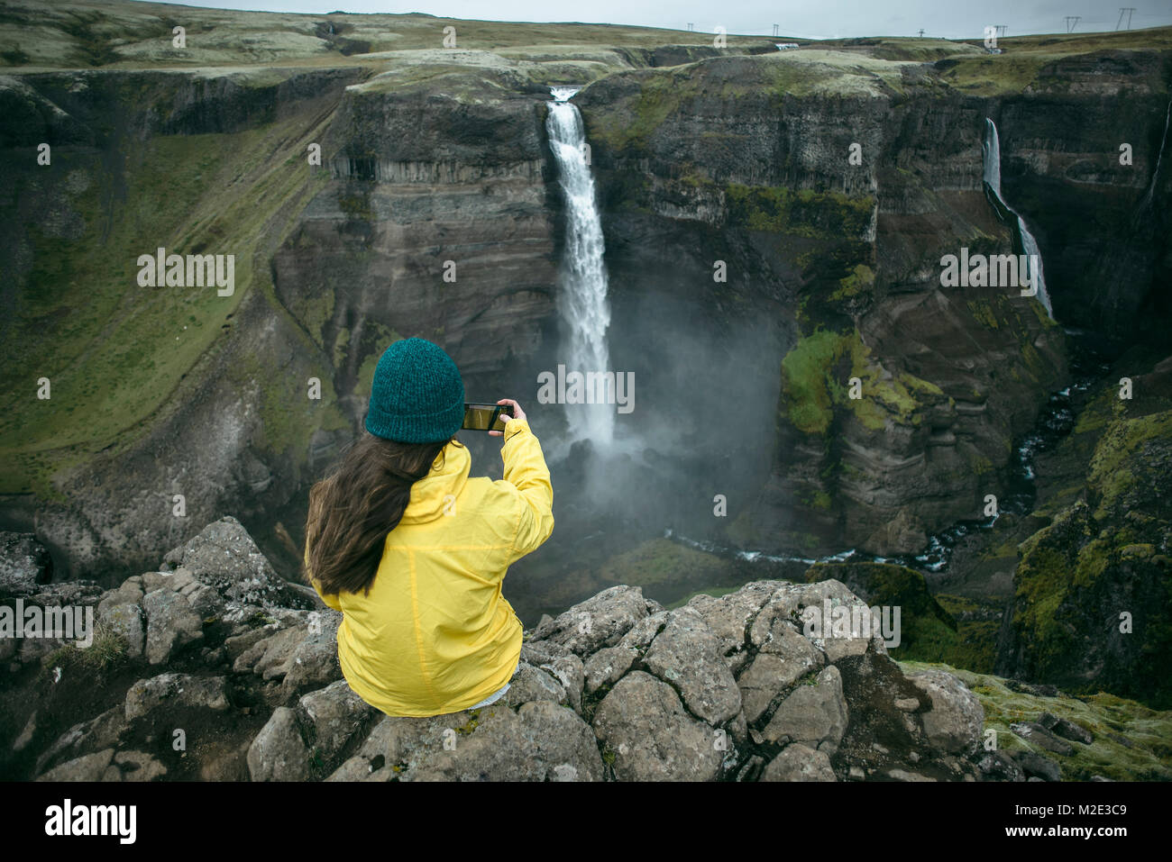 Caucasian woman sitting on cliff photographing waterfall Stock Photo