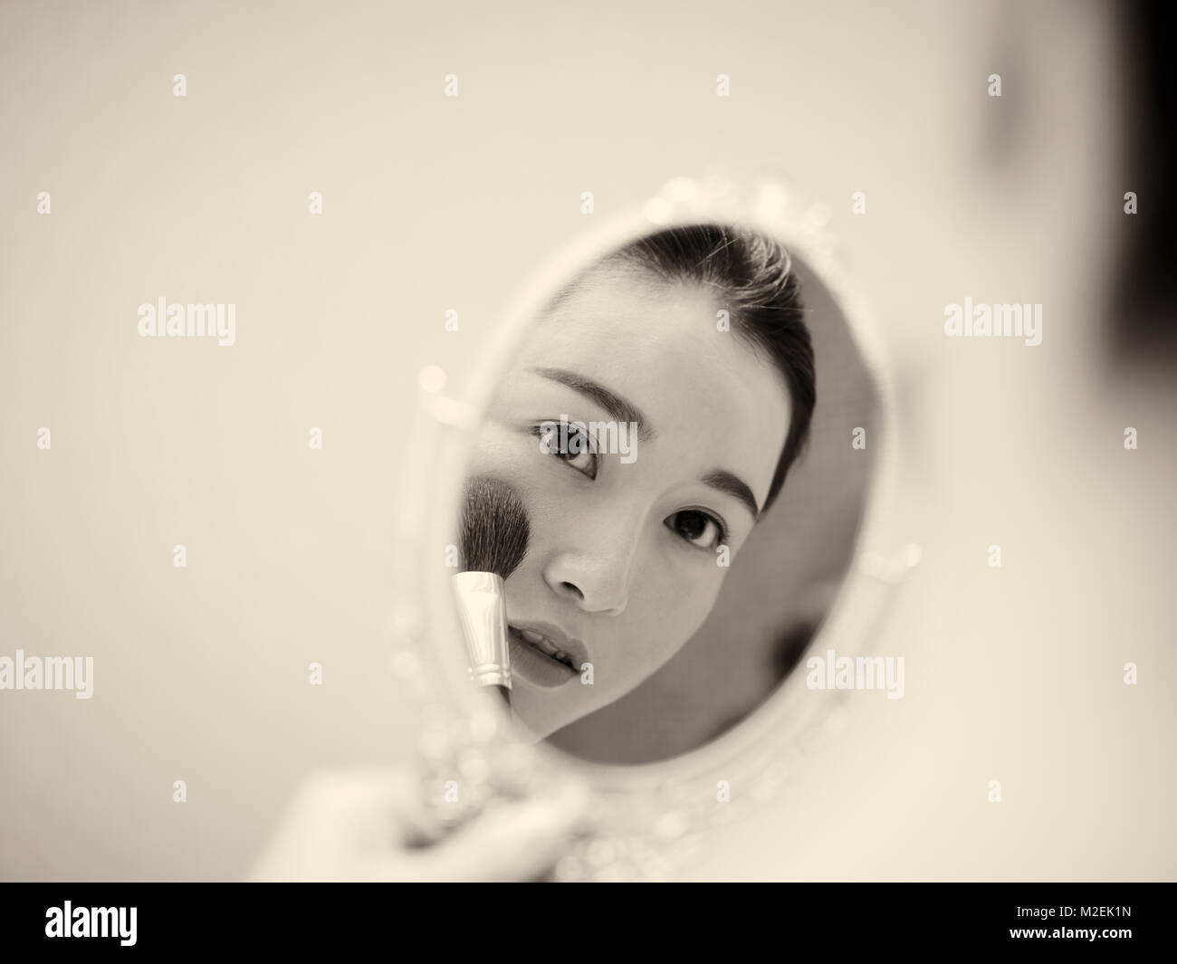 beautiful young asian woman looking at self in mirror while applying make-up on face using brush, black and white, retro style. Stock Photo