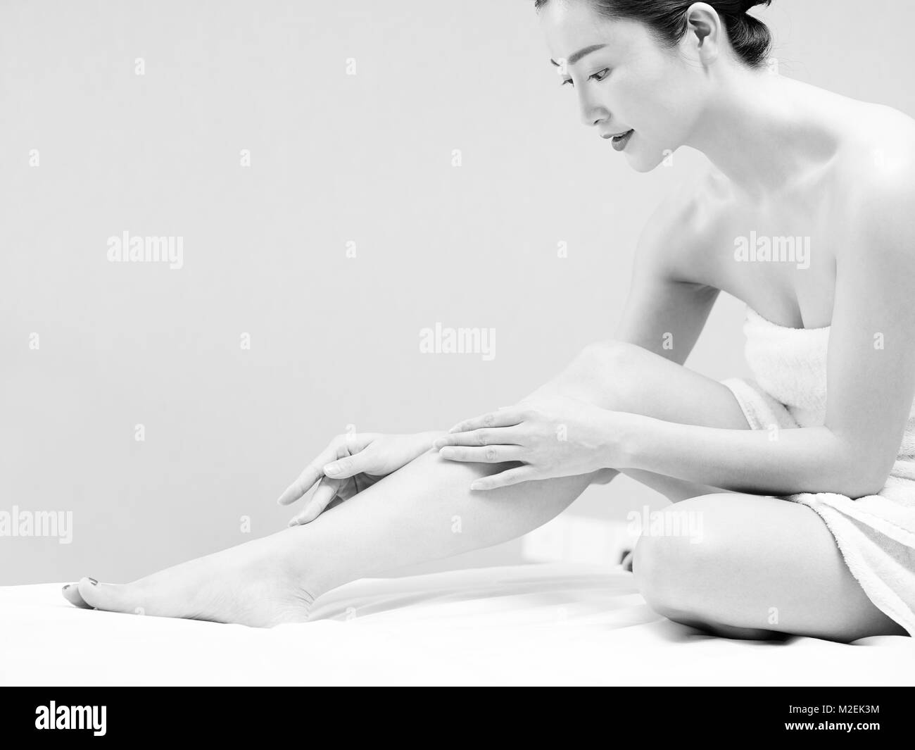 beautiful young asian woman with soft skin wrapped in white towel sitting on bed applying body lotion, black and white. Stock Photo
