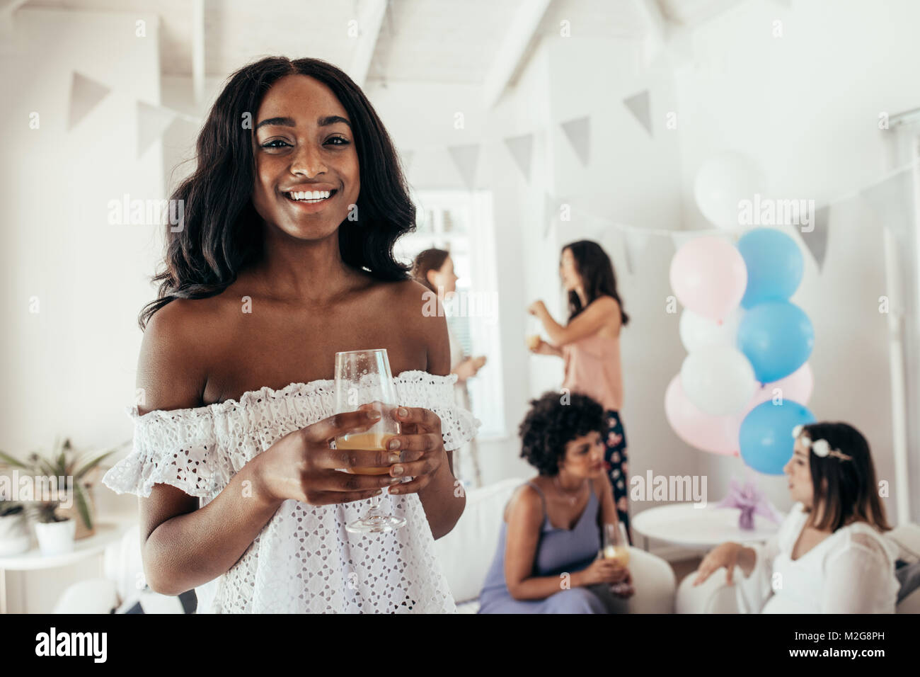 Smiling african young woman holding a glass of juice with friends in background at baby shower party. Female friends at baby shower party at home. Stock Photo