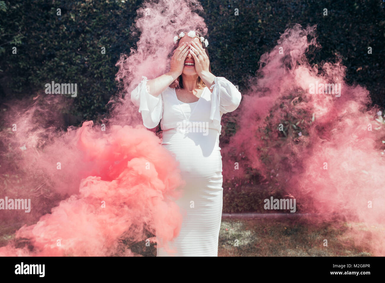 Happy pregnant woman covering her face with smoke grenade outdoors. Gender reveal party. Stock Photo