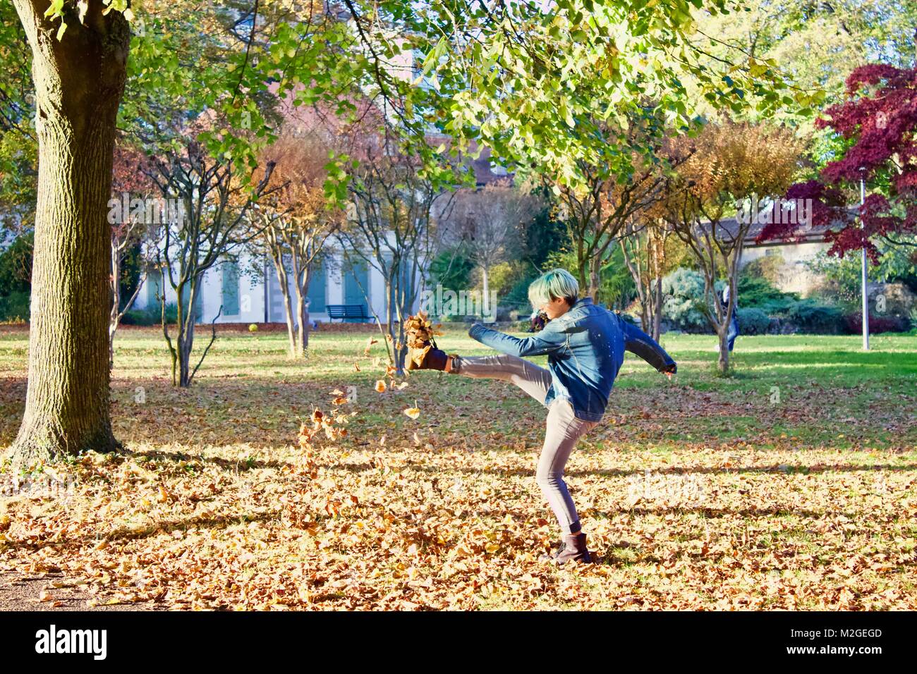 Adolescent with green hair kicking dead leaves up in the air in an empty park with lots of leaves on the ground Stock Photo