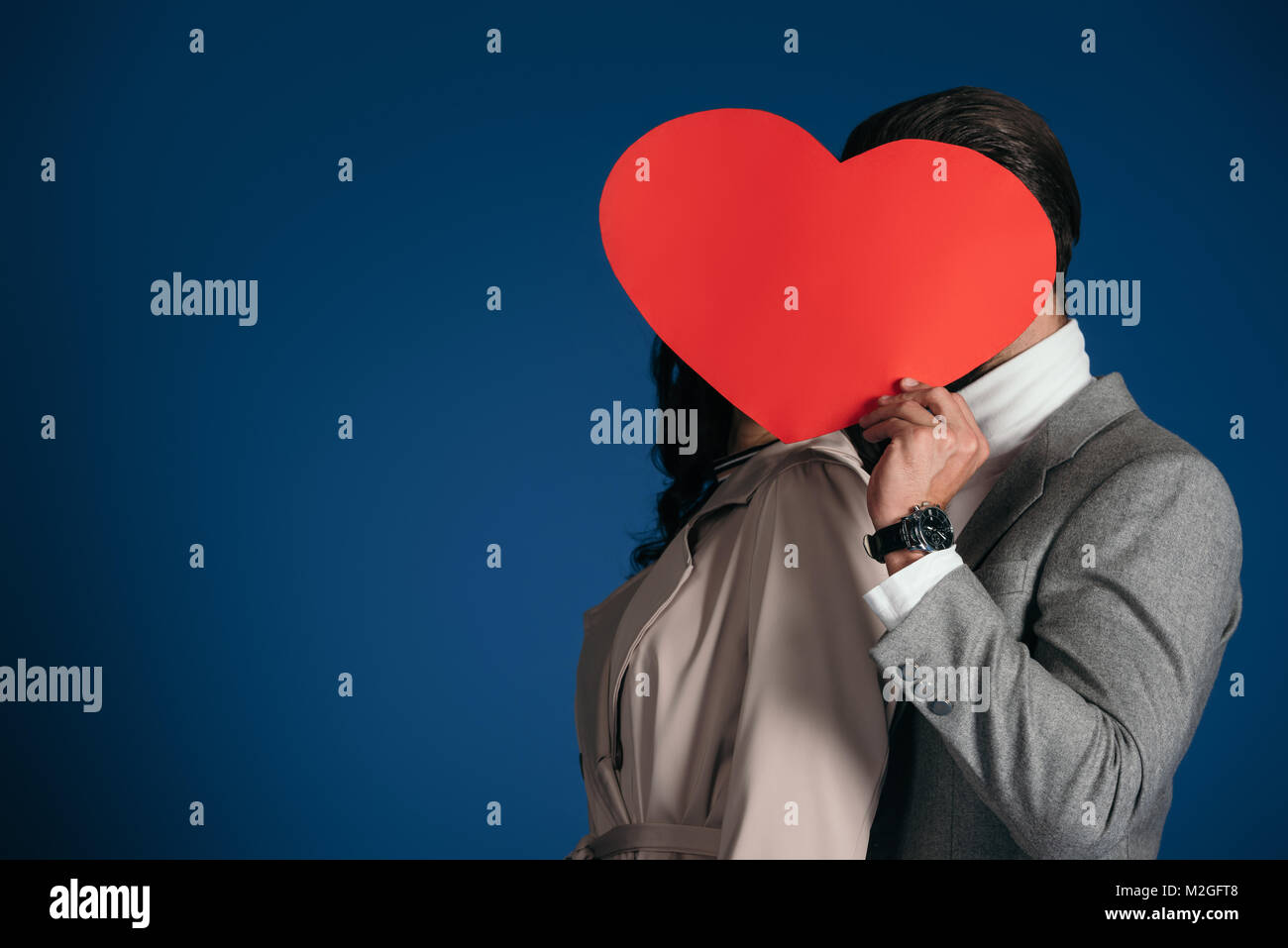 couple covering faces with heart shaped piece of paper isolated on blue