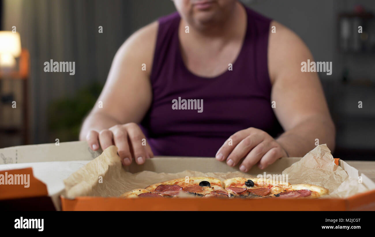 Obese man looking at fatty pizza on table, junk food addiction