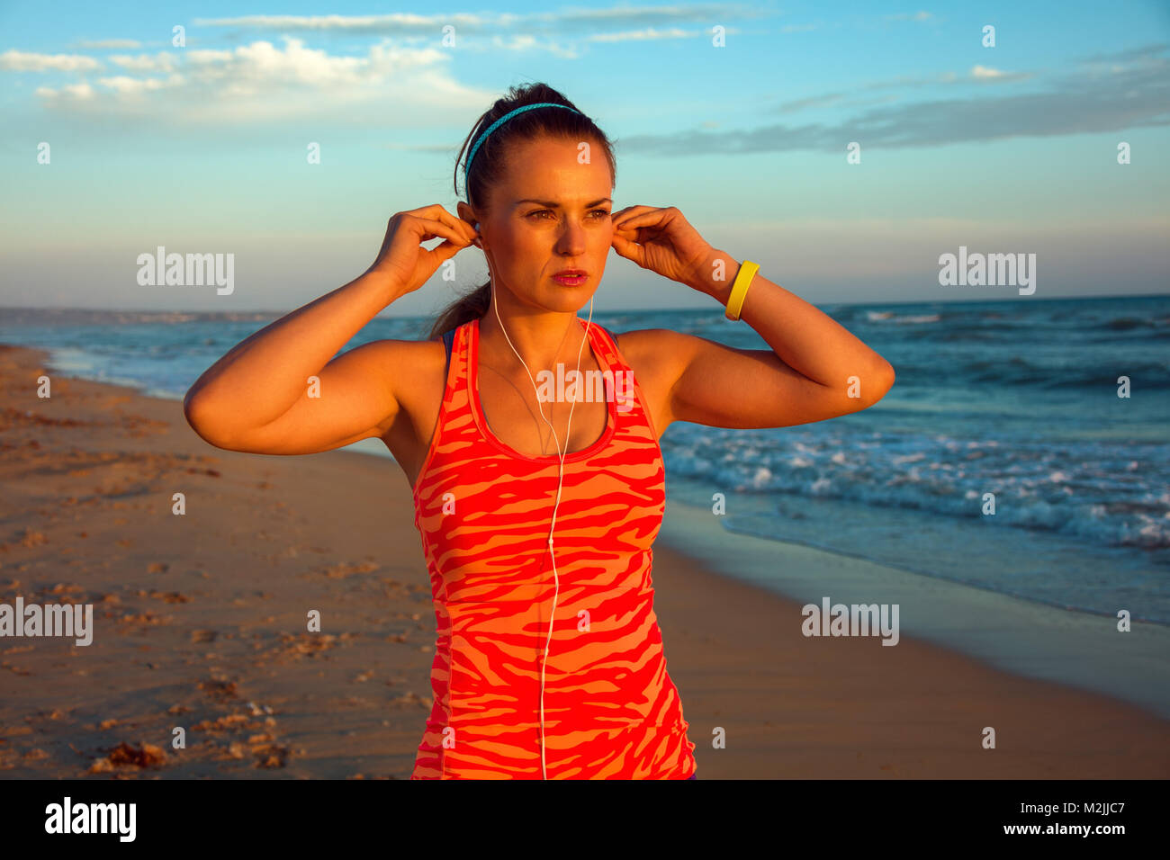 Refreshing wild sea side workout. Portrait of active woman in sportswear on the seacoast at sunset looking into the distance and listening to the musi Stock Photo