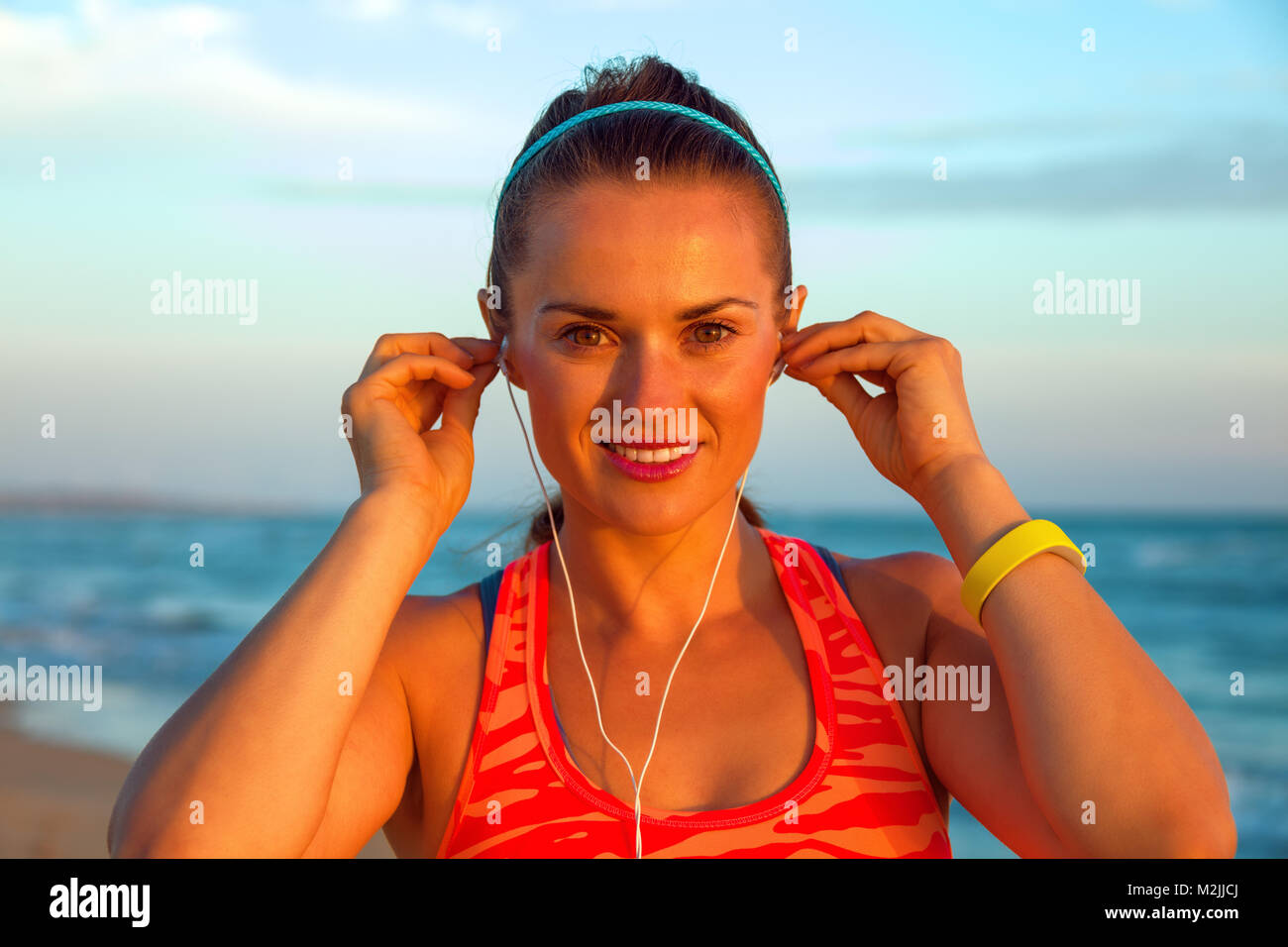 Refreshing wild sea side workout. Portrait of happy active woman in sport clothes on the seashore at sunset with headphones listening to the music Stock Photo