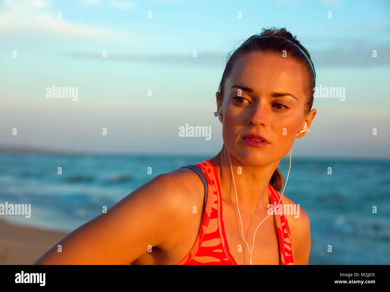 Refreshing wild sea side workout. Portrait of young woman in sport clothes on the seashore at sunset looking aside and listening to the music with hea Stock Photo