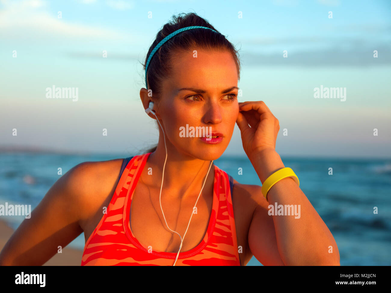 Refreshing wild sea side workout. Portrait of young woman in sportswear on the beach at sunset looking into the distance and listening to the music wi Stock Photo
