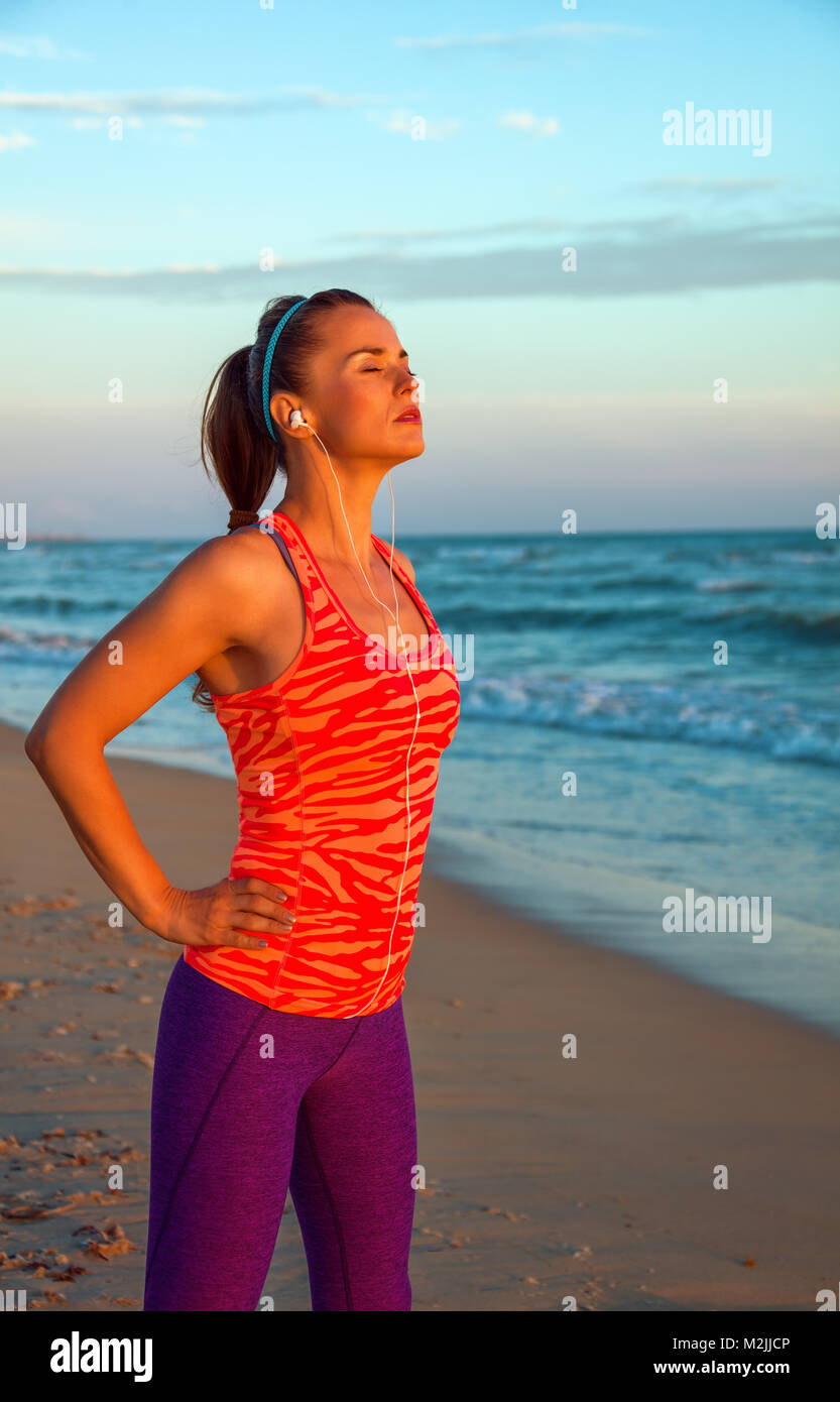 Refreshing wild sea side workout. relaxed fit woman in sports gear on the seashore at sunset with headphones listening to the music Stock Photo