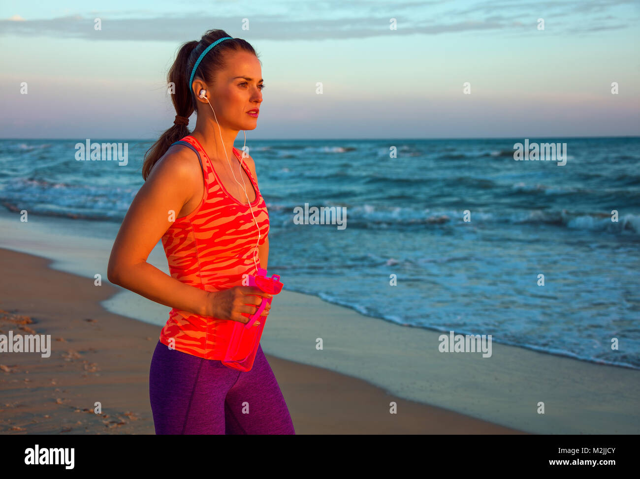 Refreshing wild sea side workout. Portrait of fit woman in sport clothes on the seashore at sunset with bottle of water looking into the distance Stock Photo