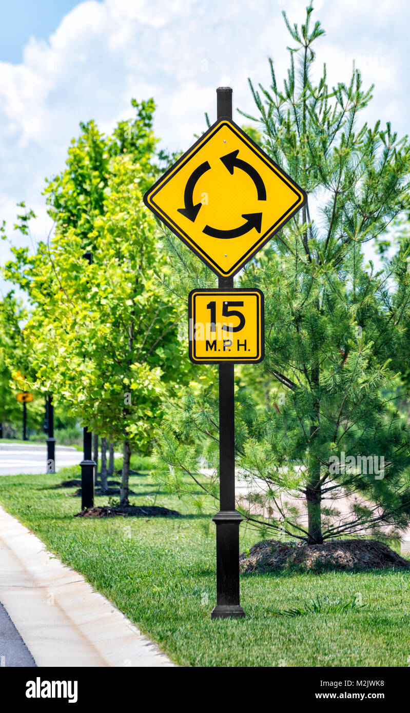 Vertical shot of a roundabout traffic sign with speed limit sign below in an upscale industrial park.  Green trees with sidewalk going past.  Cloudy s Stock Photo