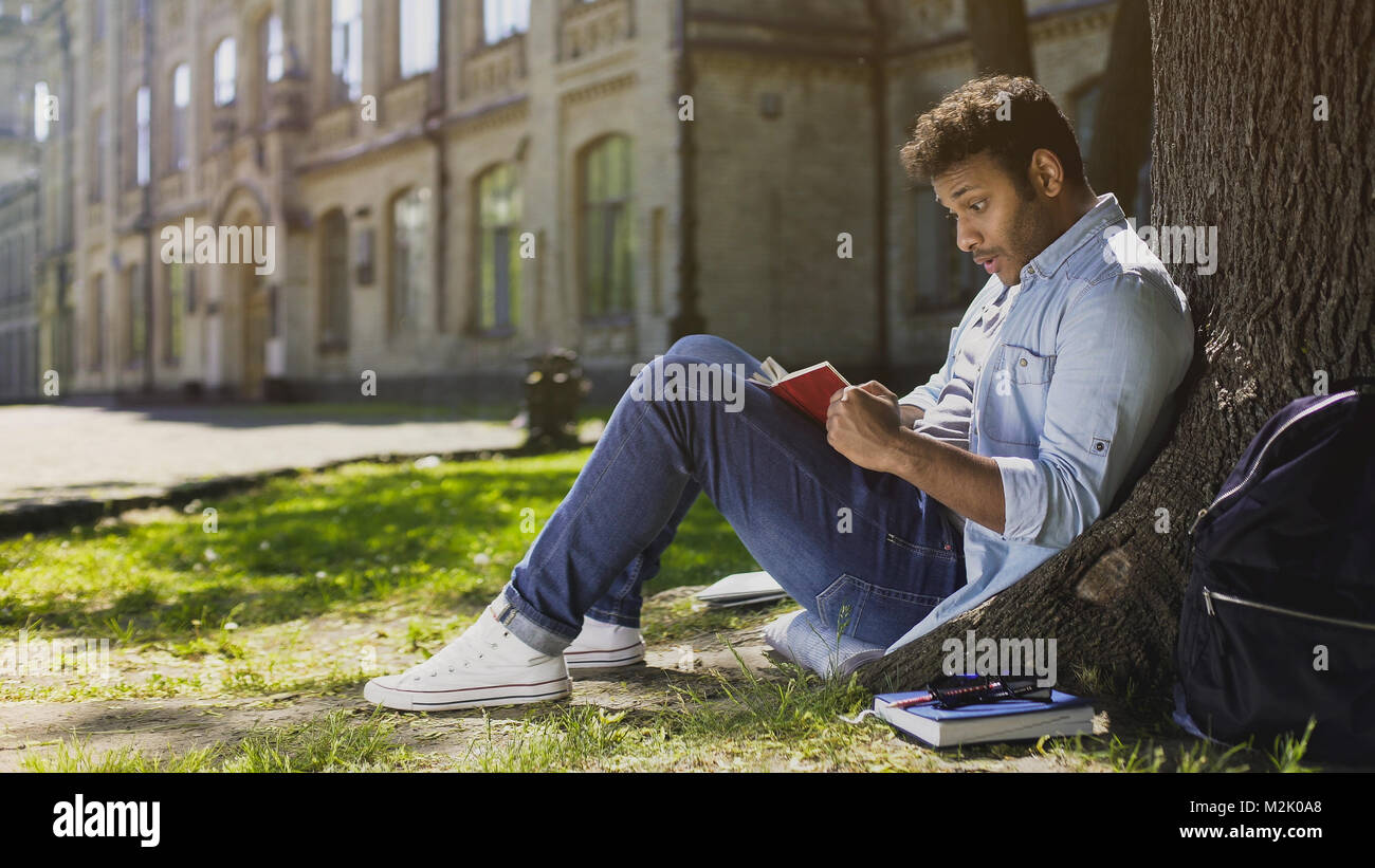 Multinational young male sitting under tree, reading gripping book, surprised Stock Photo