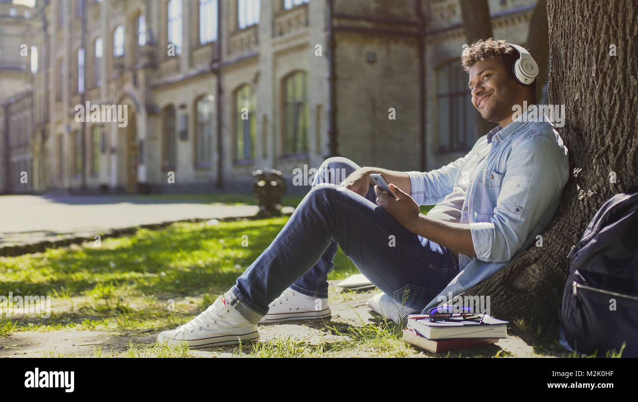 Male in headphones sitting on grass and leaning against tree, listening to music Stock Photo