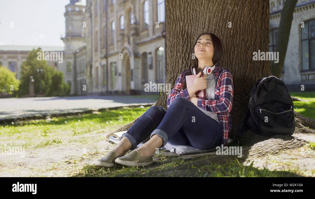 Mixed female reading book under tree, pressing it against chest, favorite book Stock Photo
