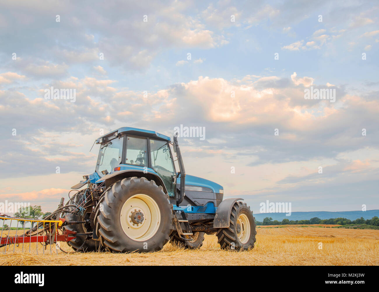 A blue tractor dominates a recently cut hayfield against the backdrop of a blue sky and clouds. Catoctin are mountains in the distance. Stock Photo