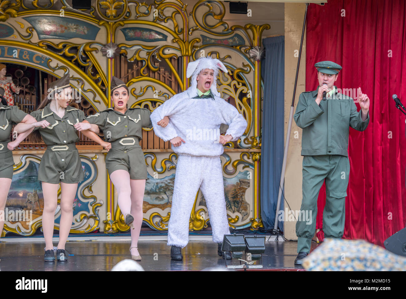 Female dancers in military attire link arms with male dancer in sheep costume to do the cancan while a man sings, Masham, North Yorkshire, UK Stock Photo