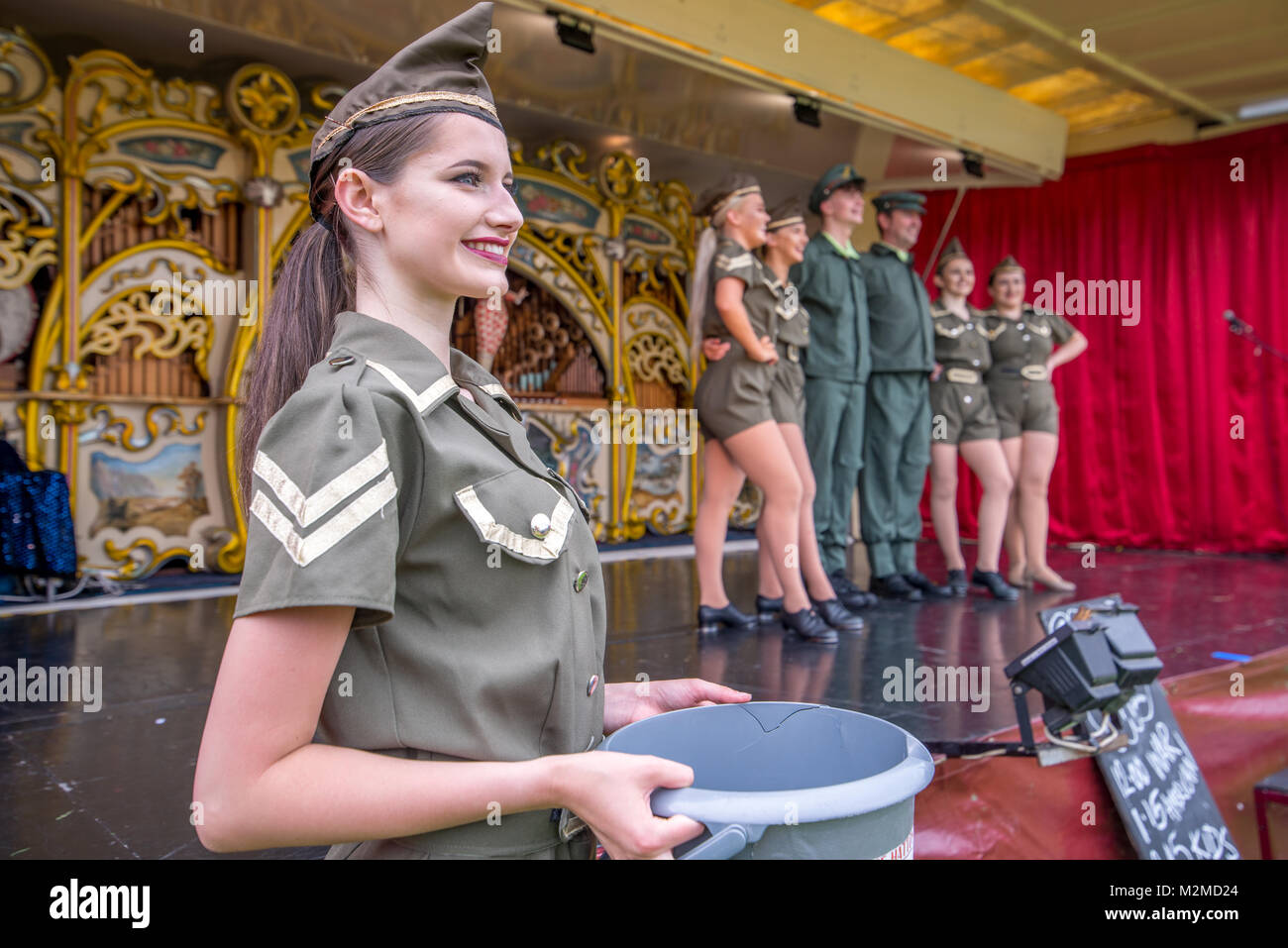 Close up of female dancer in military outfit holding collection bucket while her fellow dancers stand on top of fairground organ, Masham, North Yorksh Stock Photo