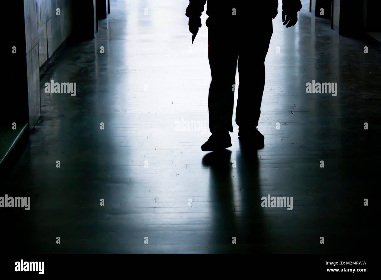 Blurry trap in dark alley in the night, silhouette of a man from waist down holding sharp object in hand while approaching Stock Photo