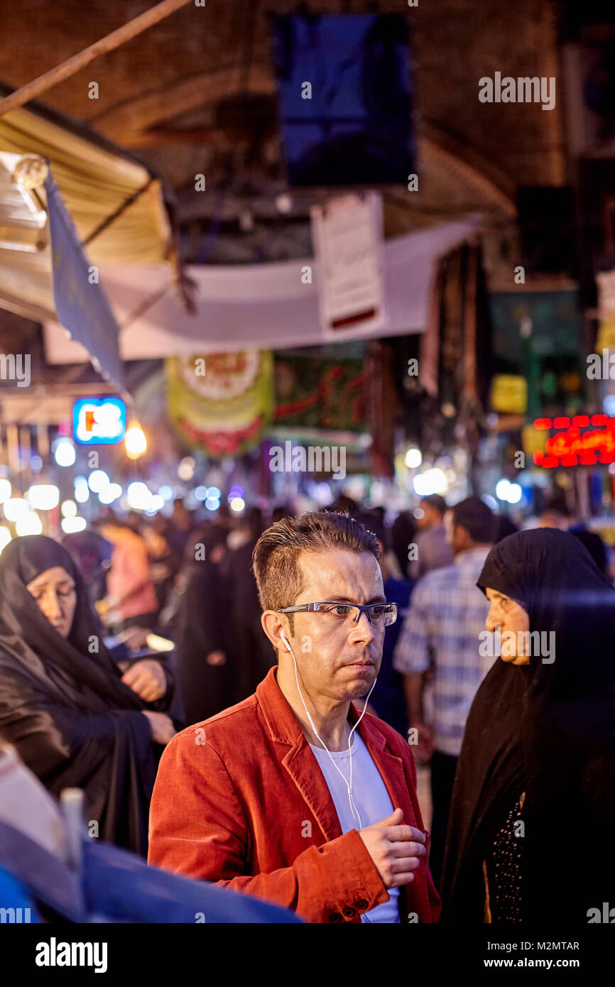 Tehran, Iran - April 27, 2017: An Iranian man with glasses, in-ear headphones, and in a red velvet jacket surrounded by muslim women in a black chador Stock Photo