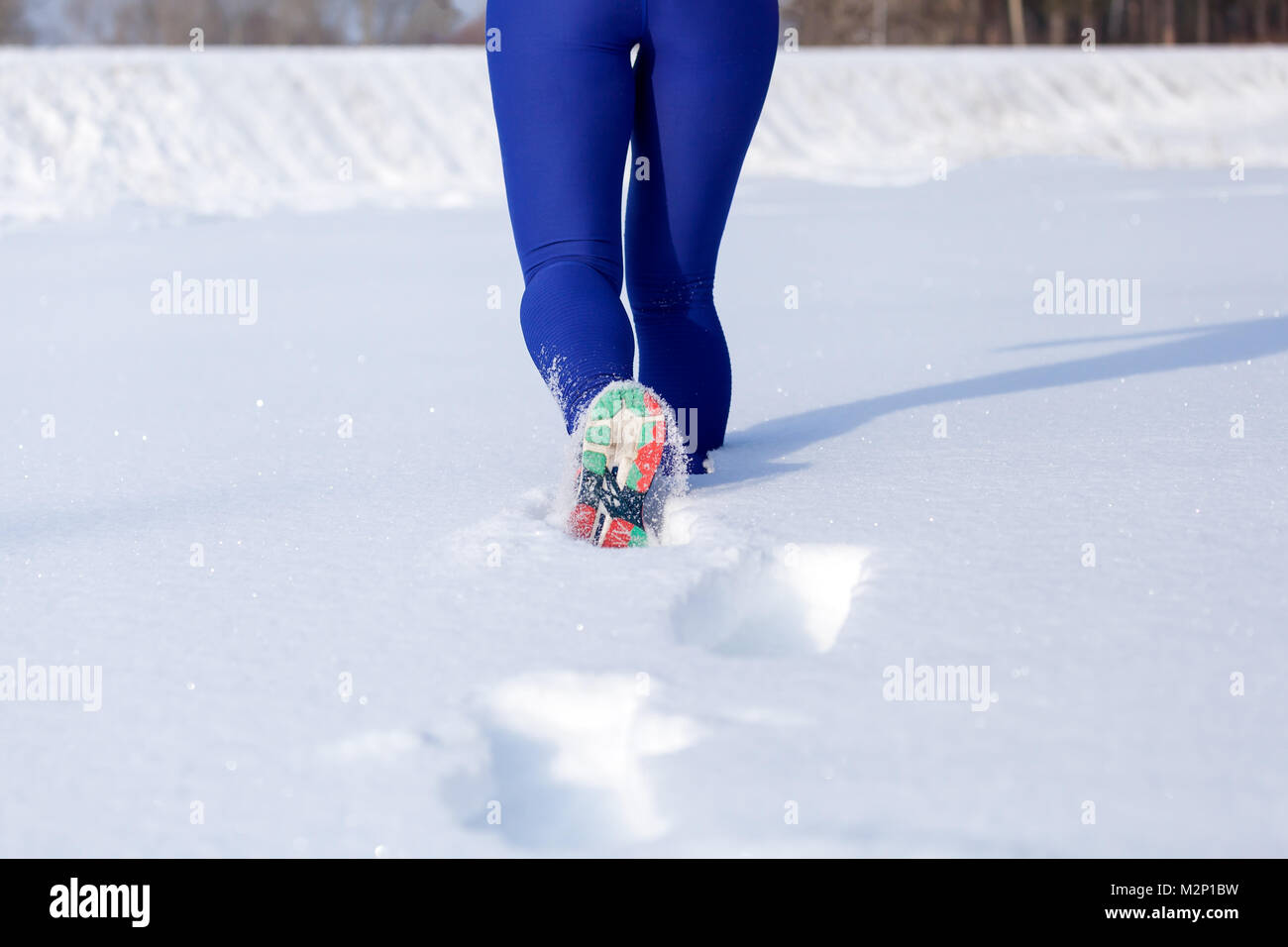 Close-up of a young woman in bright purple leggings and sneakers running through the winter snow on a bright winter day, rear view Stock Photo