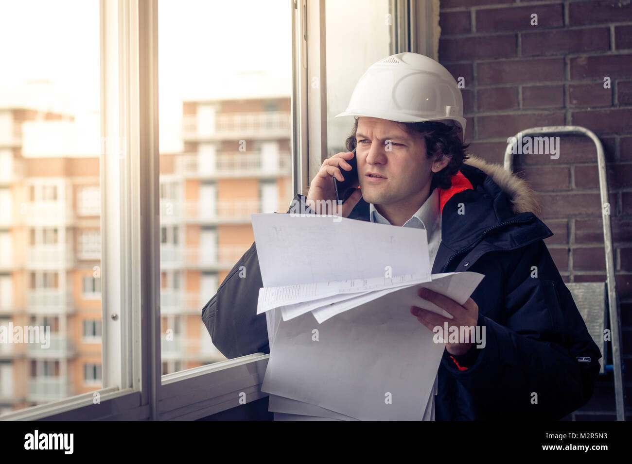 Portrait of concerned unshaven builder telling by mobile while keeping different projects in hand. Communication and work concept. Soft focus, toned Stock Photo