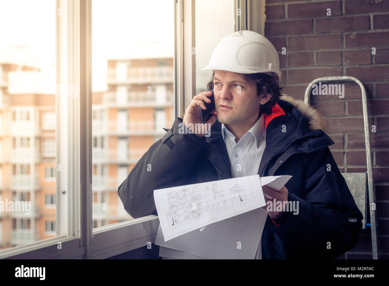 Portrait of concerned unshaven builder telling by mobile while keeping different projects in hand. Communication and work concept. Soft focus, toned Stock Photo
