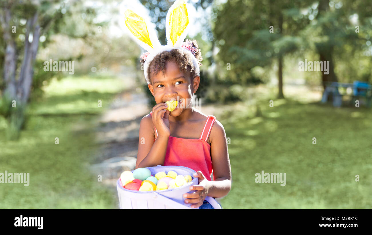 Cute and beautiful African American girl wearing bunny ears, eats with joy Easter marshmallow candy while holding Easter basket full of eggs. Girl sit Stock Photo