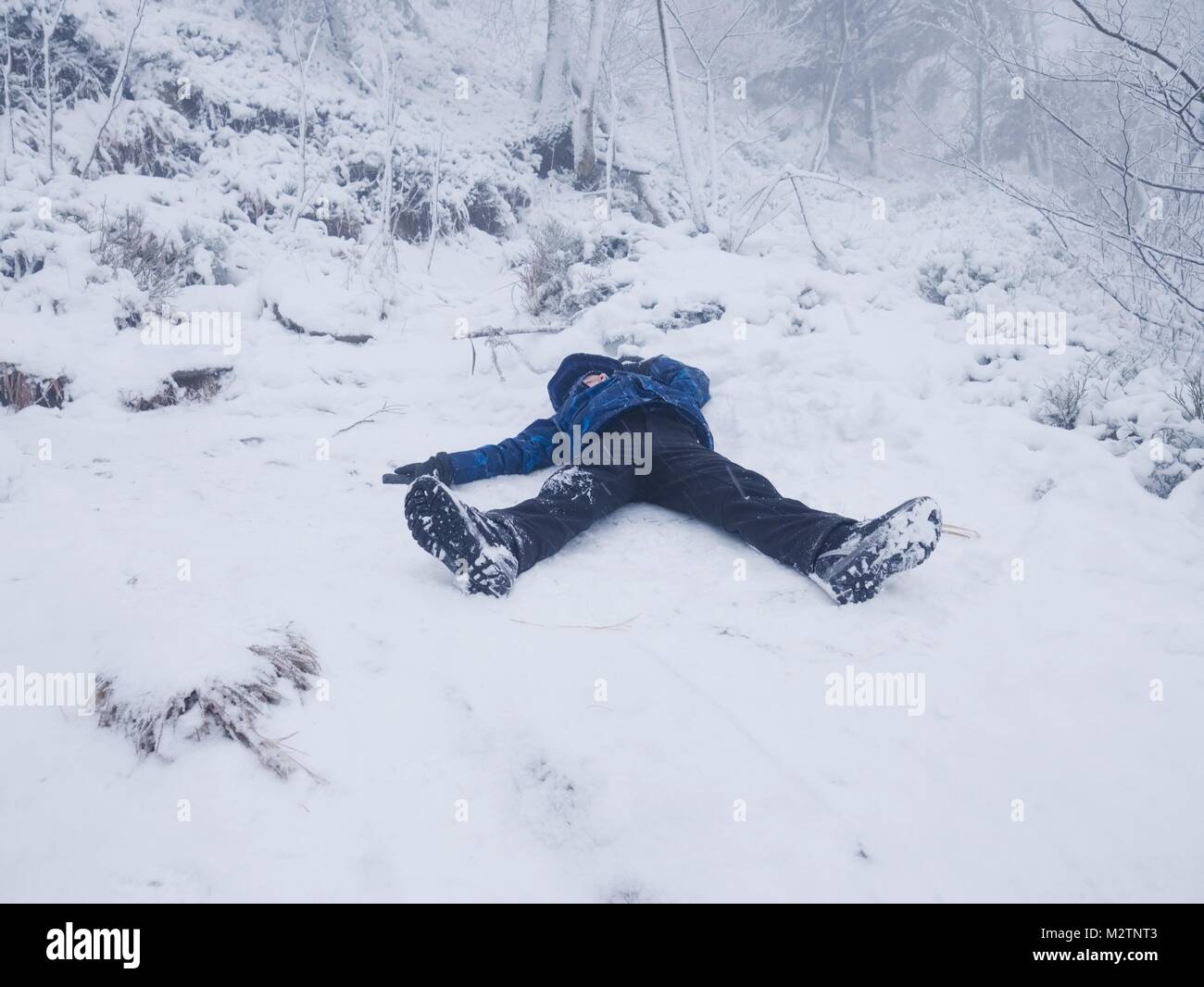 Funny boy laying in snow and makes an angel with spread arms. Kid play game in fresh snow on stony hill. Stock Photo