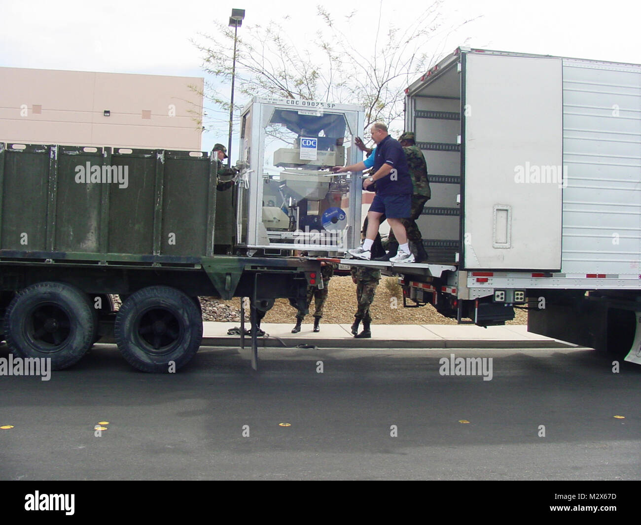 This image, was captured at an undisclosed location, and depicts a Strategic National Stockpile (SNS) 12-hour Push-Package being unloaded off of a transport truck, and on to a military transport vehicle, as a number of people oversaw this materiel transfer procedure. It appears that this group was composed of SNS staff, and military personnel. It appeared that this module contained what might have been a radiographic device. These modular containers are ready-made for shipment, while still at the SNS warehouse. This module had yet to be be locked to its metal palette. Stock Photo