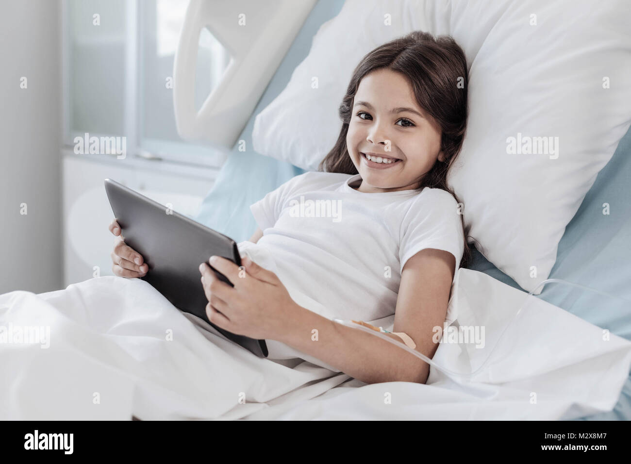 Brave girl playing on touchpad while lying in hospital bed Stock Photo