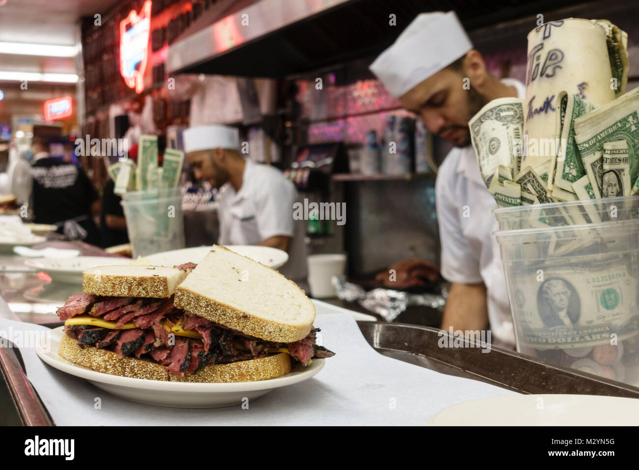 Hard-working staff making pastrami sandwiches in the open kitchen at Katz's Delicatessen, a famous New York City restaurant. Tips in cup on counter. Stock Photo