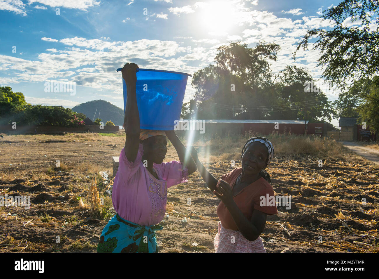 Woman lifting a bucket of water on the head of another woman, Lake