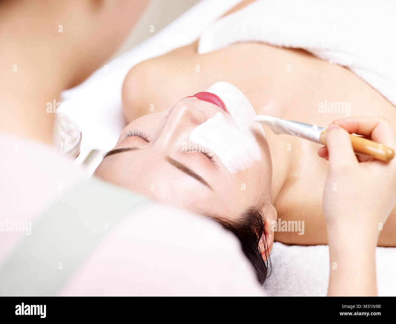 asian beautician applying white facial mask with a brush on face of a young woman. Stock Photo